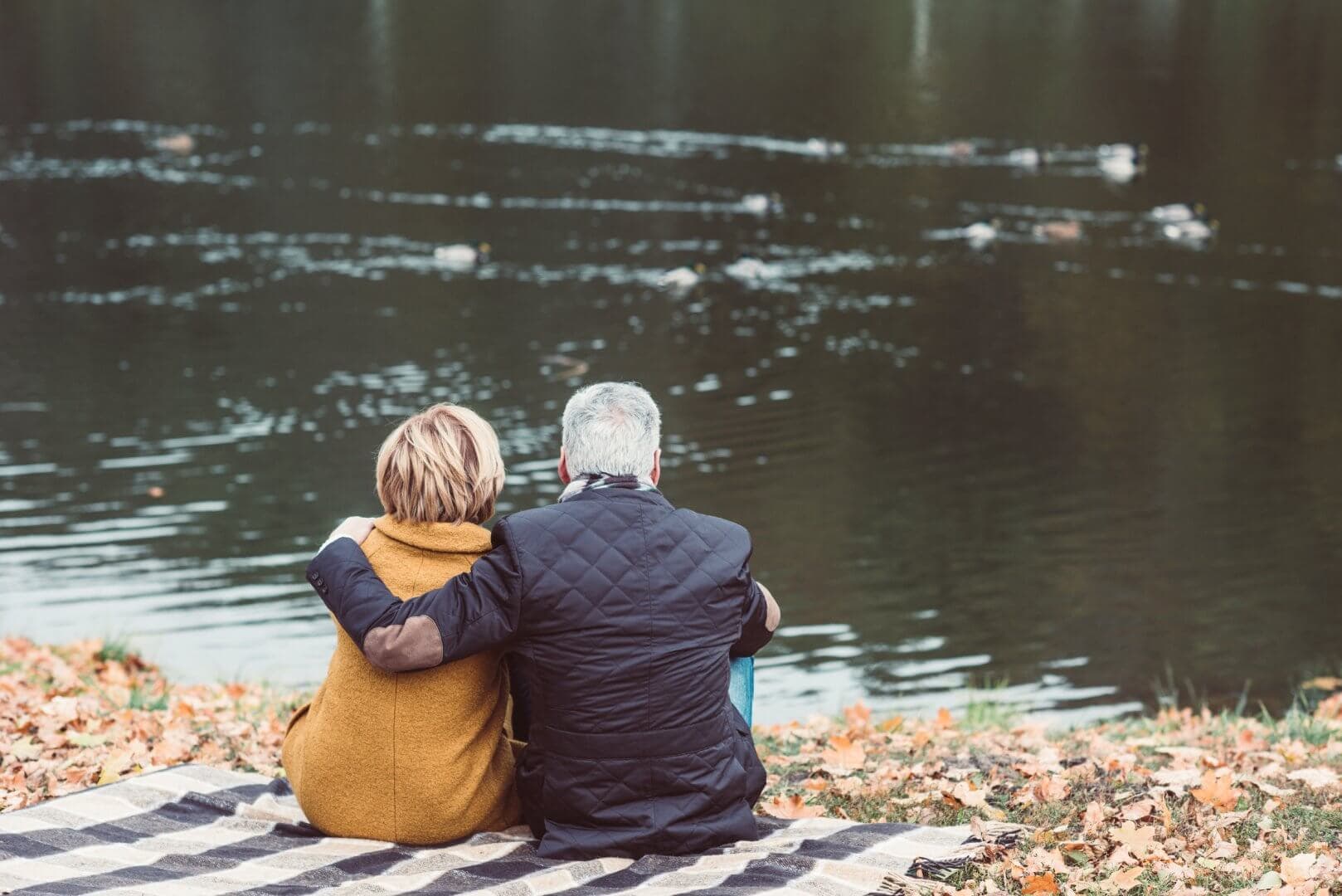 A couple sits on a blanket by a lake, with their backs to the camera, surrounded by autumn leaves. - Home Instead Southampton