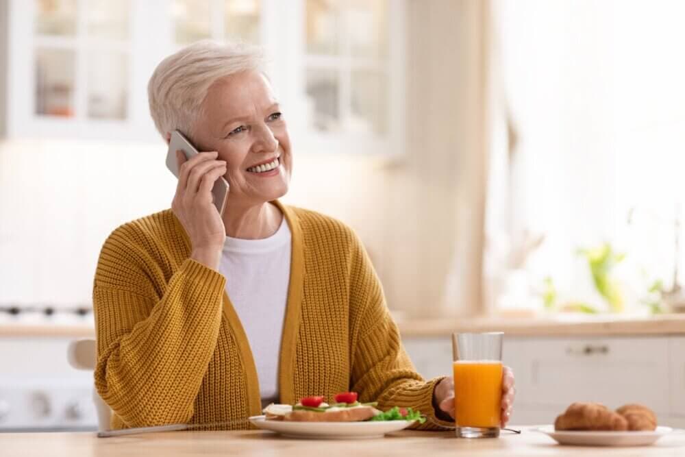 A smiling woman with short white hair, in a yellow cardigan, talks on the phone while holding a glass of orange juice. - Home Instead