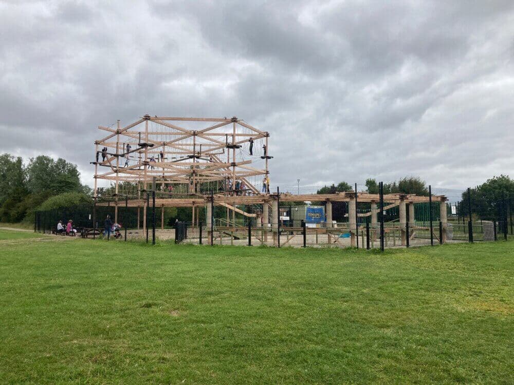 Outdoor adventure park with a tall wooden ropes course structure surrounded by grass and trees under a cloudy sky. - Home Instead