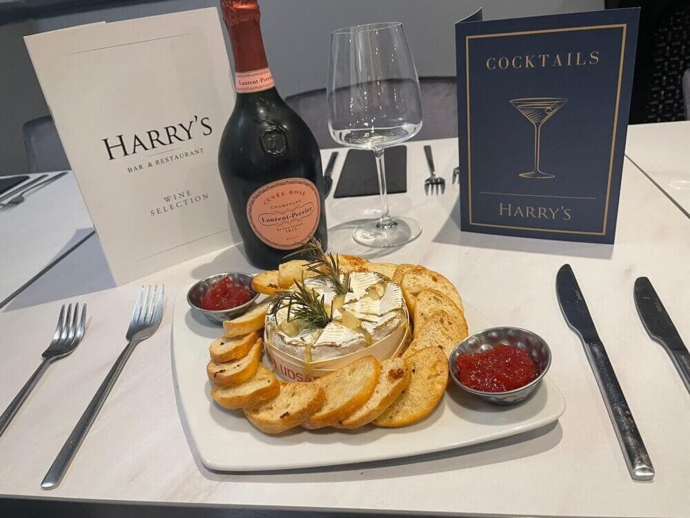 Baked camembert with bread slices, jam, a bottle of Laurent-Perrier, and menus on a restaurant table. - Home Instead