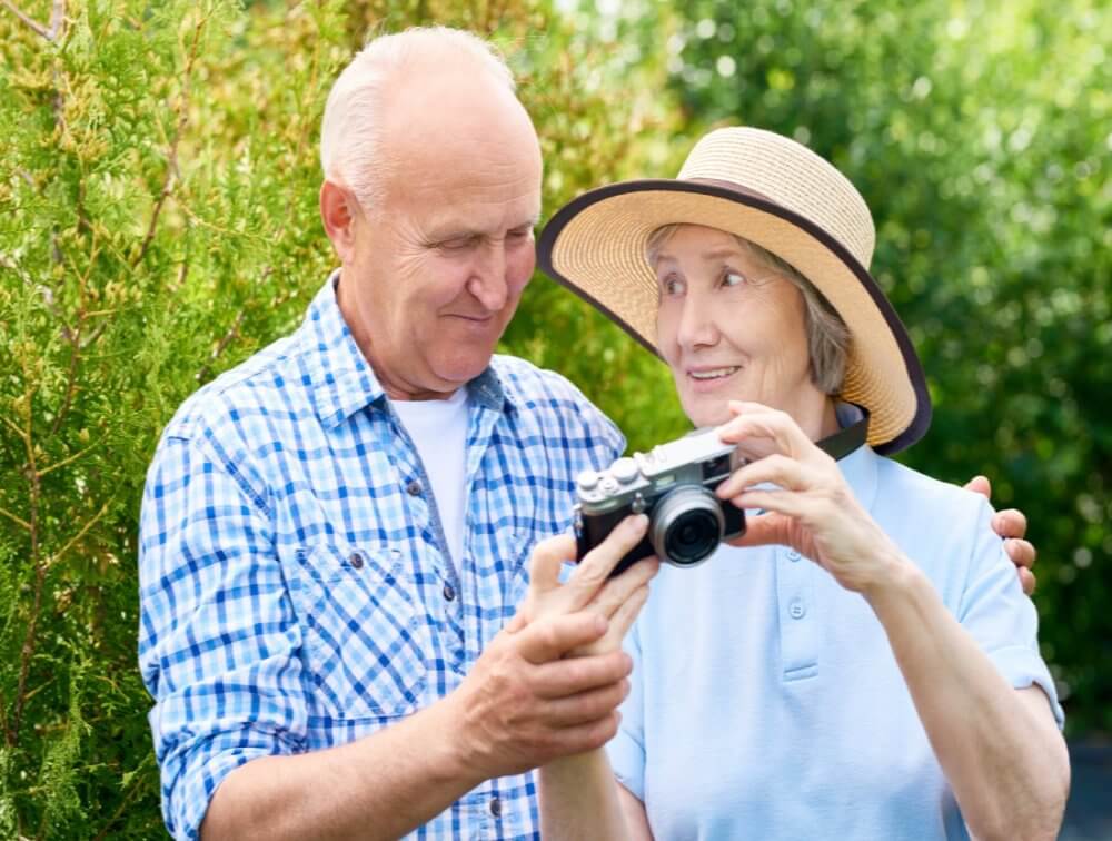 Elderly couple with a camera outdoors. The man in plaid shirt holds the woman in a sunhat, pointing at the camera screen. - Home Instead