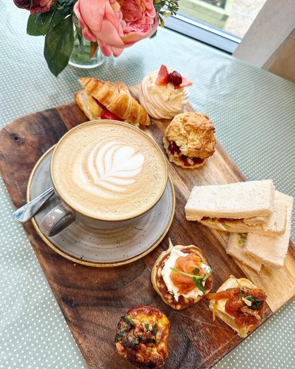 A latte with leaf art is surrounded by assorted pastries, sandwiches, and a floral arrangement on a wooden board. - Home Instead