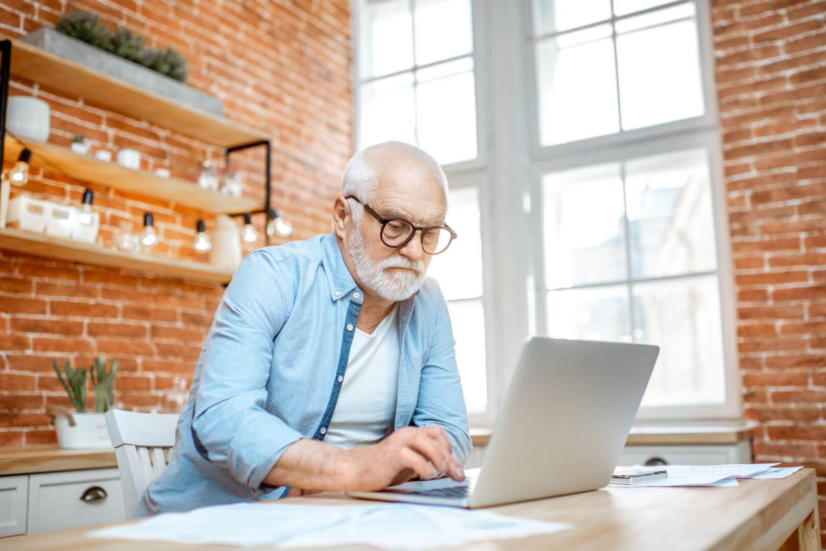Elderly man with glasses working on a laptop in a bright room with brick walls and large windows. - Home Instead