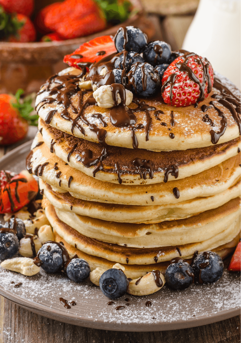 A stack of pancakes topped with strawberries, blueberries, nuts, chocolate drizzle, and a dusting of powdered sugar. - Home Instead