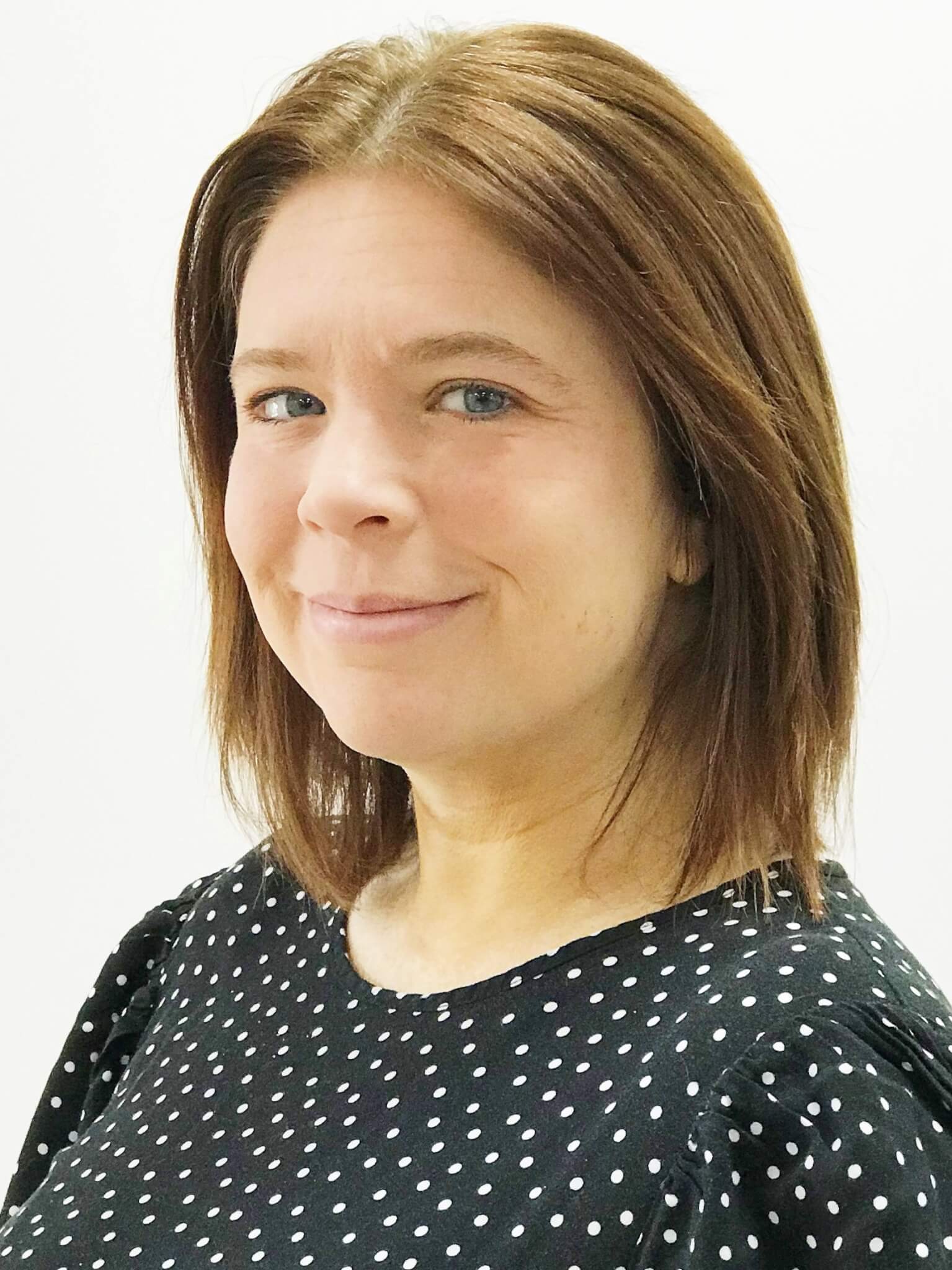 Smiling person with shoulder-length brown hair wearing a black shirt with white polka dots against a white background. - Home Instead