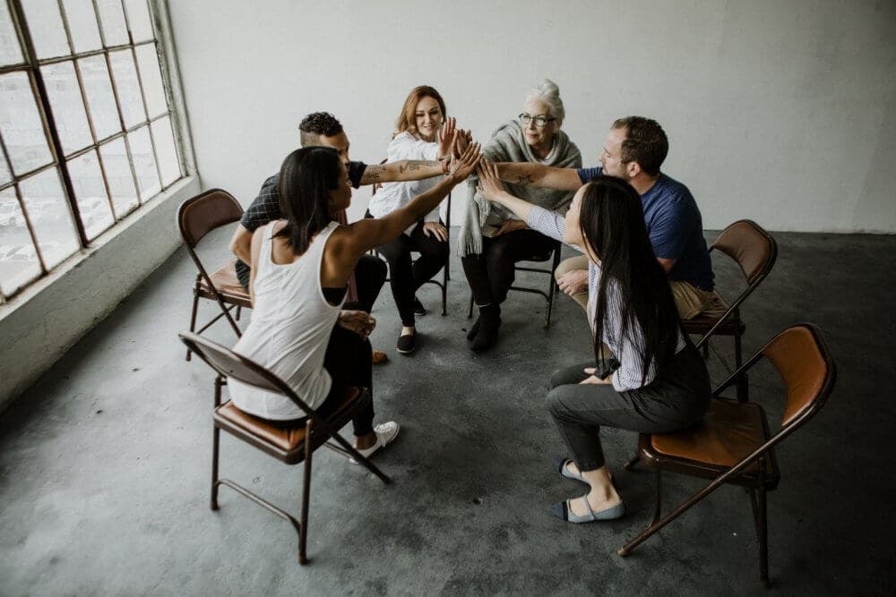 A group of six people sitting in a circle in a loft, reaching towards the center for a group high-five. - Home Instead