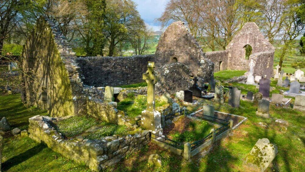 Ruins of an old stone church surrounded by gravestones and trees, located in a lush green landscape. - Home Instead