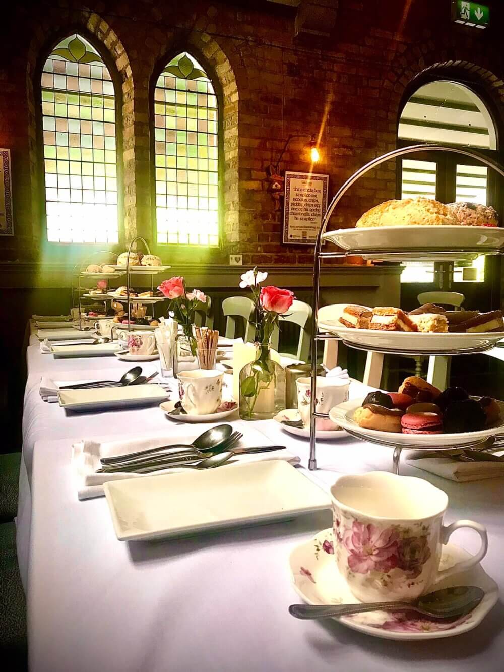 A table set for an afternoon tea with floral china, tiered trays of pastries, and pink roses in a sunlit room with stained glass windows. - Home Instead