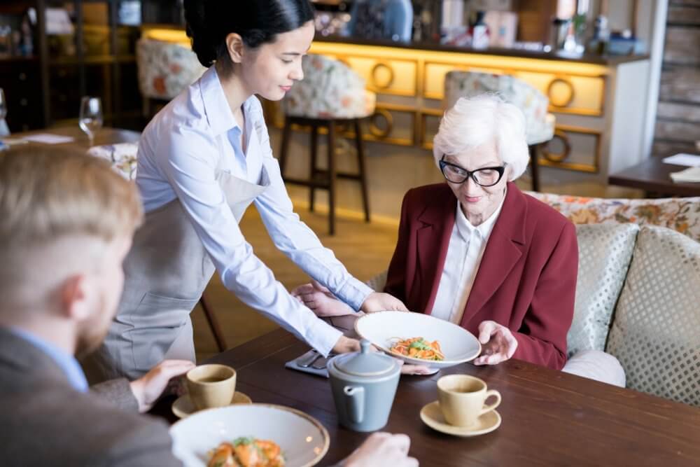 Waitress serving food to an elderly woman in a red blazer at a restaurant, with a man sitting nearby. - Home Instead