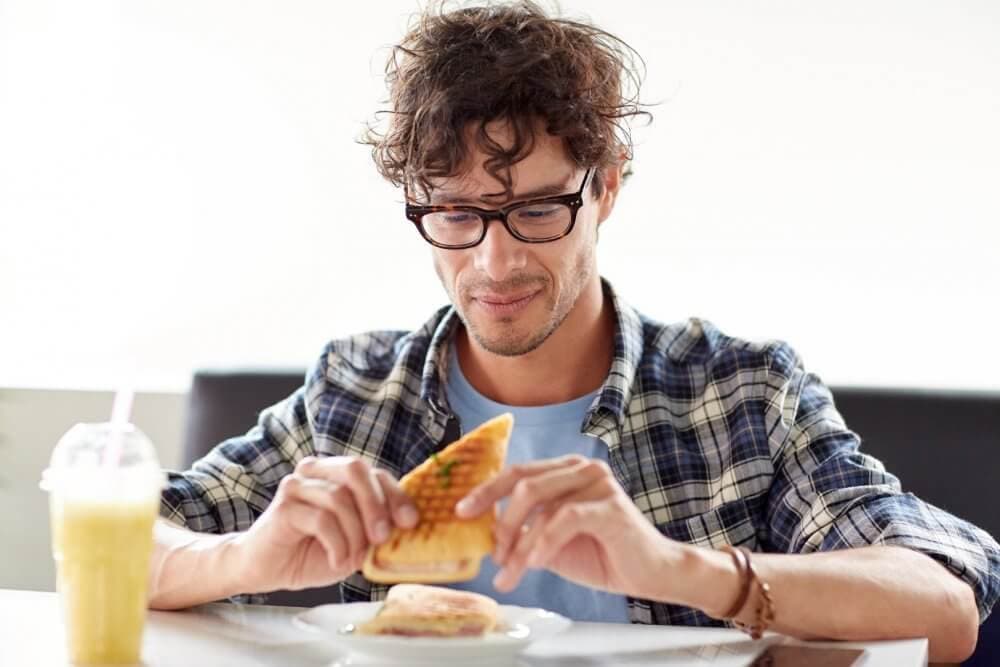 A man with glasses and curly hair smiles while eating a panini, with a smoothie on the table. - Home Instead