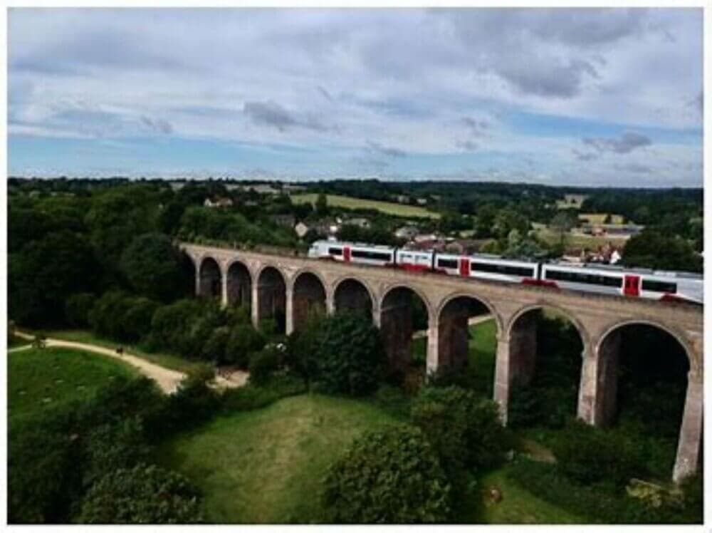 A train crosses a tall viaduct bridge over a lush green landscape with trees, fields, and scattered buildings. - Home Instead