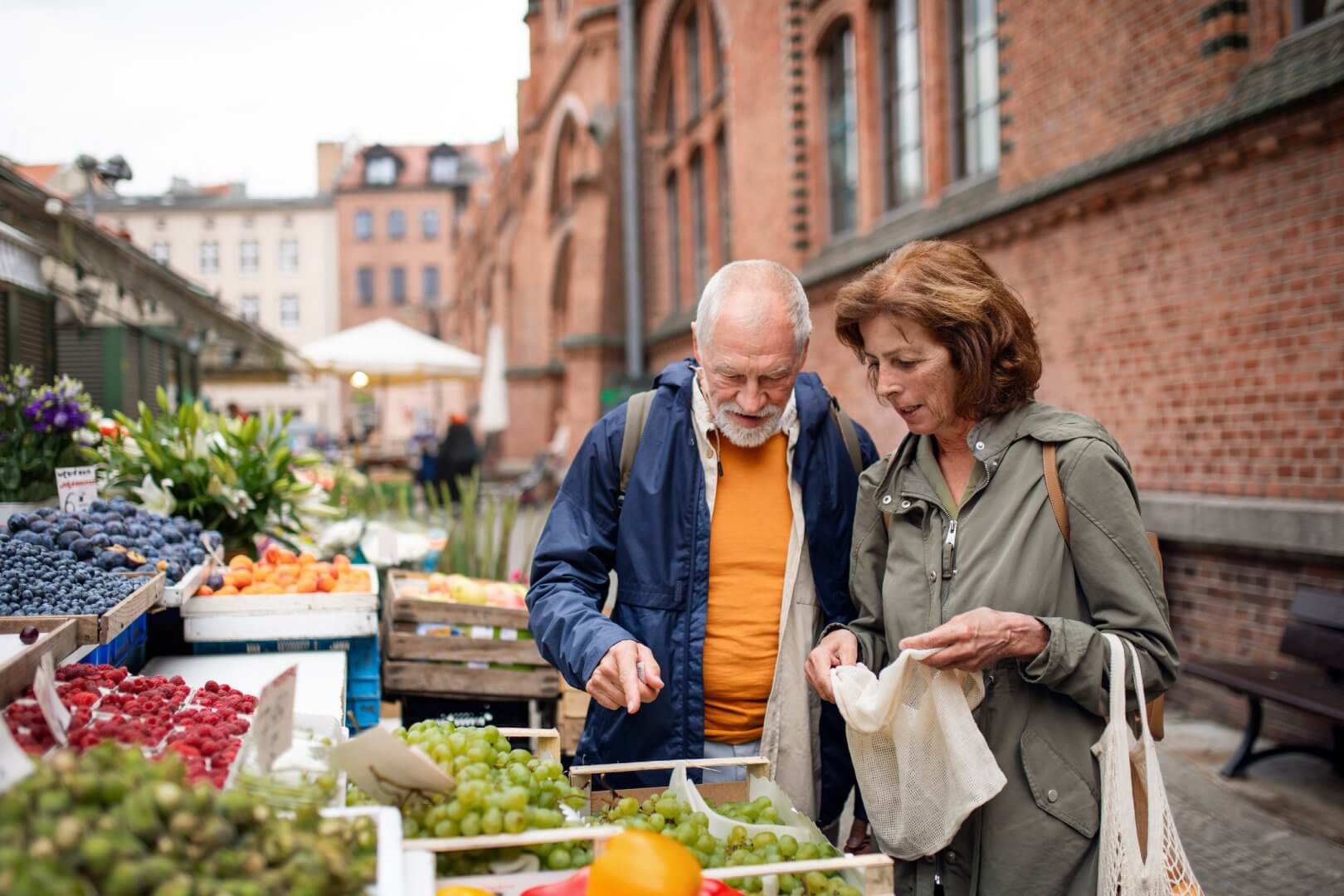 An older couple shops for fruit at an outdoor market, standing by a produce stall with grapes, berries, and other fruits. - Home Instead