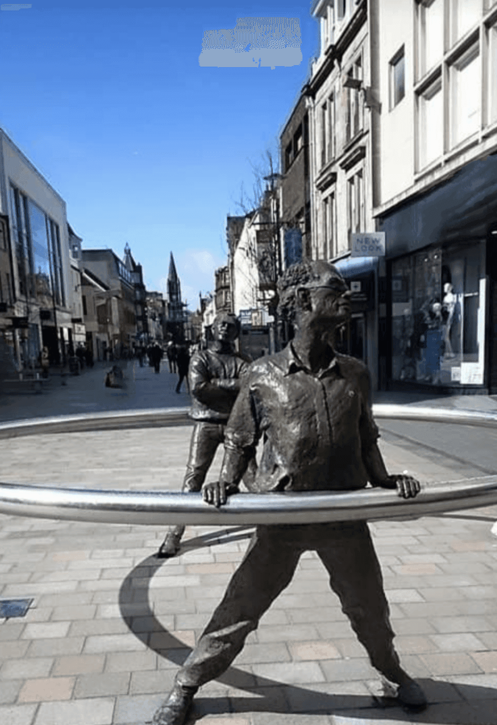 Two bronze statues of men standing with large hoops in a pedestrian shopping street on a sunny day. - Home Instead