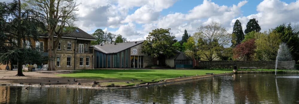 A serene park features a modern building, lush greenery, a pond with ducks, a fountain, and trees under a partly cloudy sky. - Home Instead