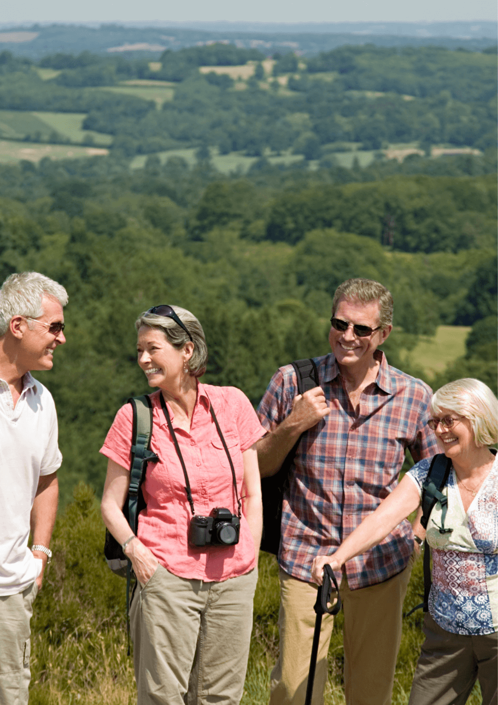 Four smiling adults with backpacks and walking gear stand on a grassy hill with a scenic landscape in the background. - Home Instead