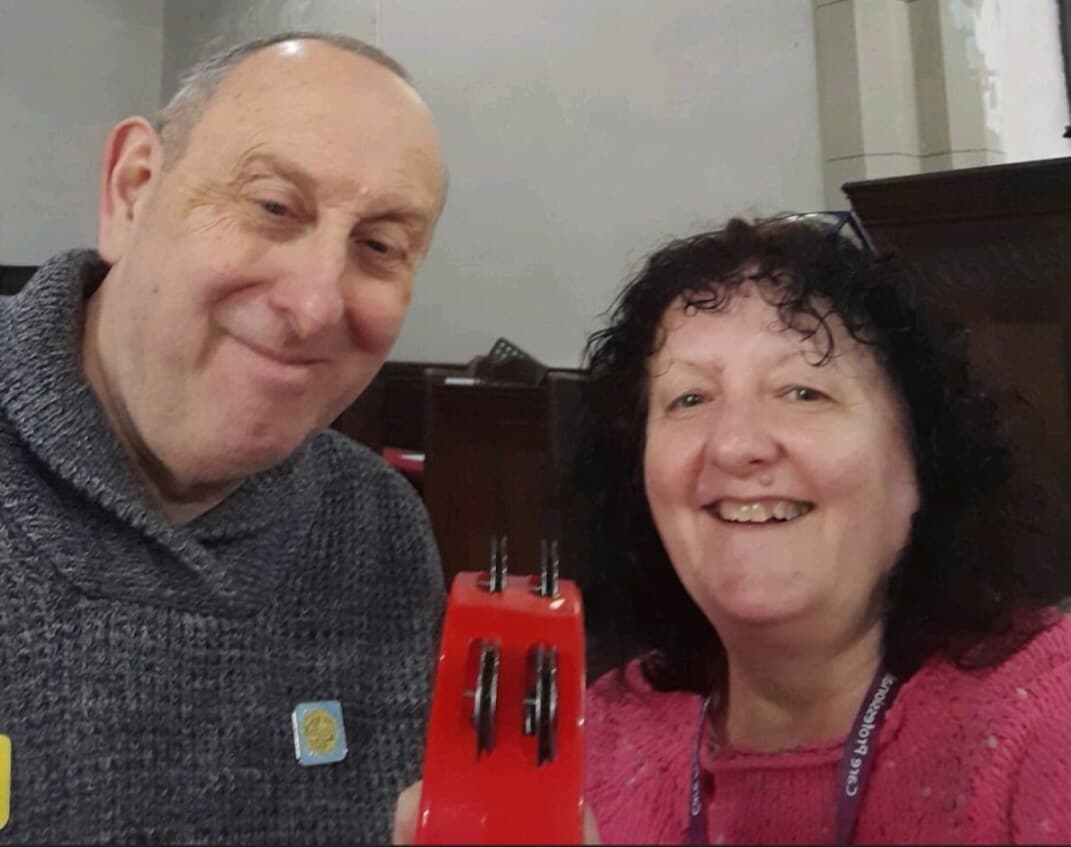 Two people smiling indoors holding a red object with three small wheels. - Home Instead