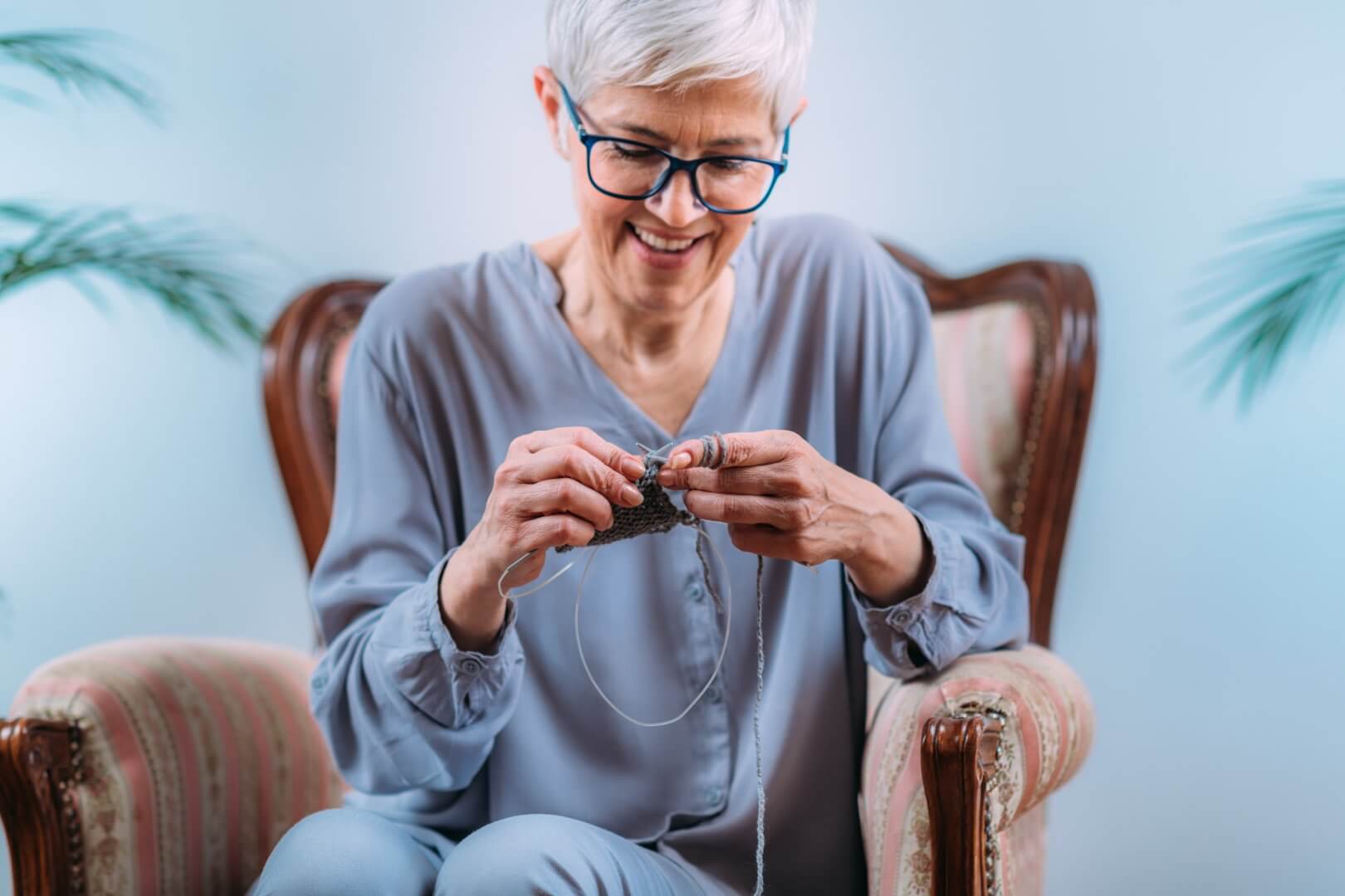 An elderly woman with glasses sits in an armchair, smiling while knitting. - Home Instead