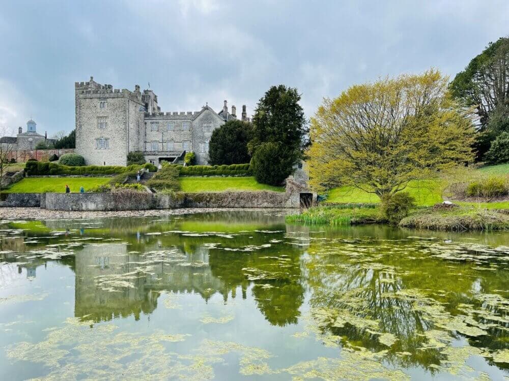 A castle overlooks a pond with greenery and yellow tree reflections under a cloudy sky. - Home Instead