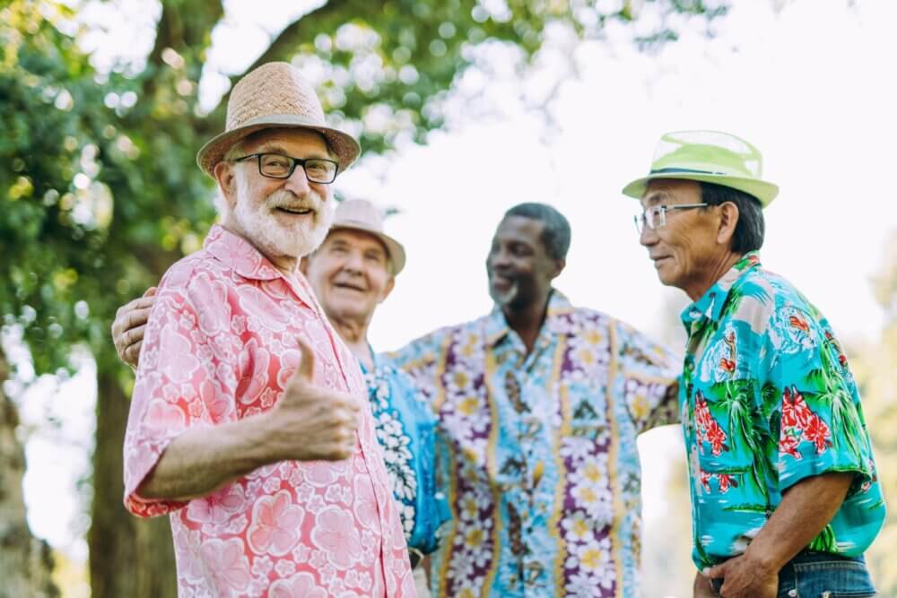 Group of four senior men in colorful shirts and hats, smiling and chatting outdoors. One man is giving a thumbs-up. - Home Instead