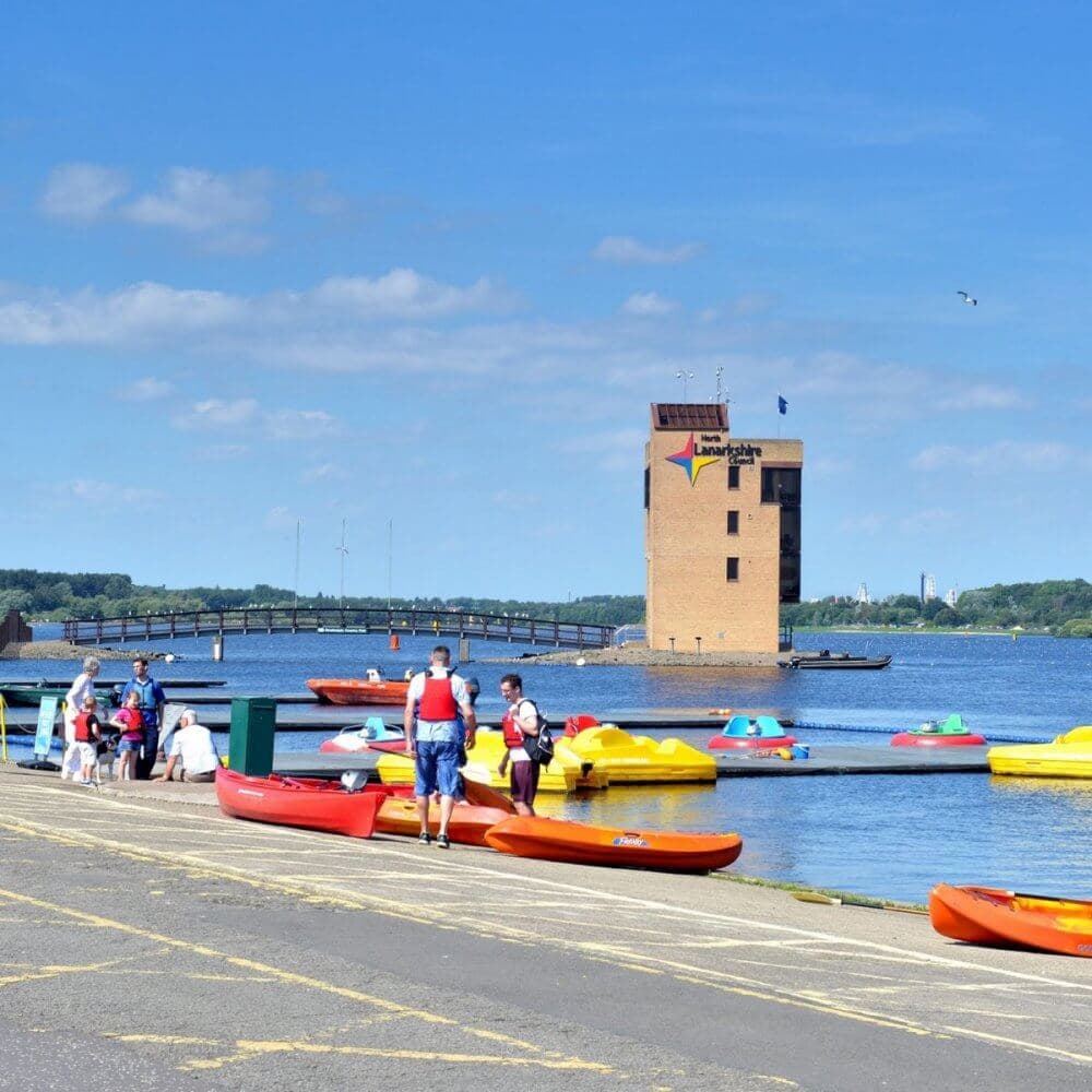 People by the dock with colorful kayaks and paddle boats on a sunny day, waterside building in the background. - Home Instead