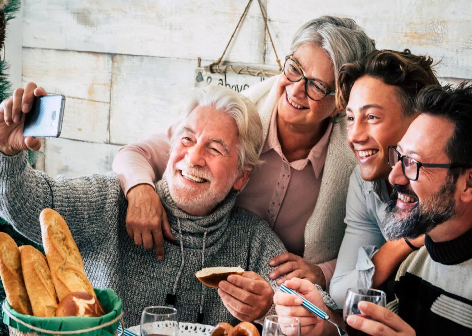 Four people smiling and taking a selfie at a dining table with bread and drinks. - Home Instead