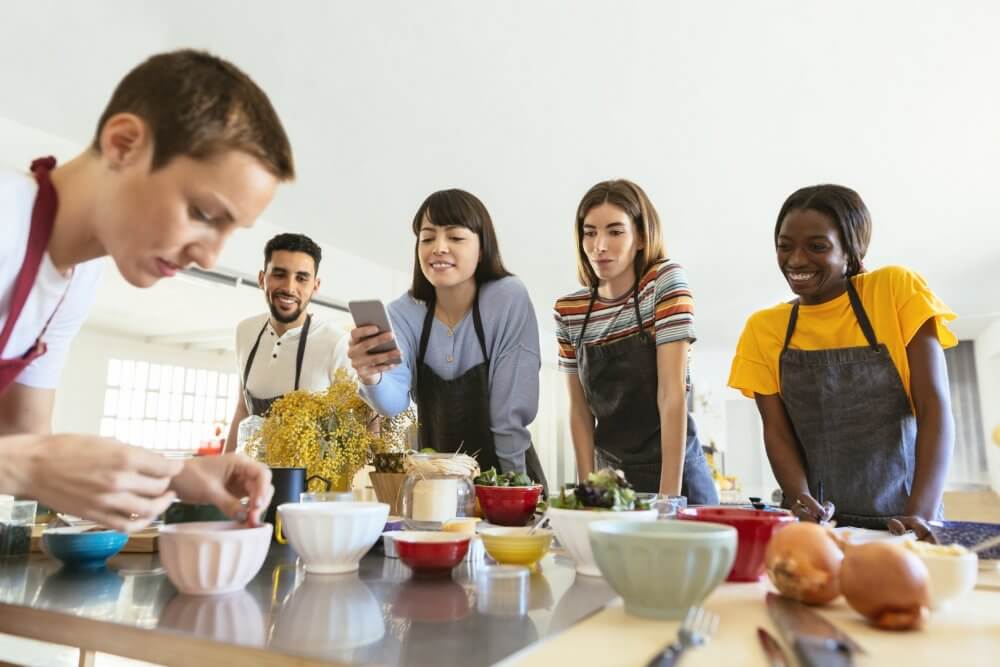 Five people in aprons are gathered around a counter, observing someone prepare food in a brightly lit kitchen. - Home Instead