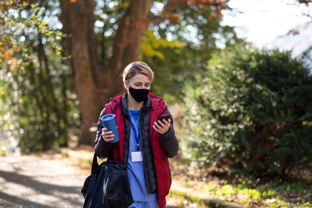 Person wearing a mask, holding a drink and looking at their phone while walking outside among trees. - Home Instead