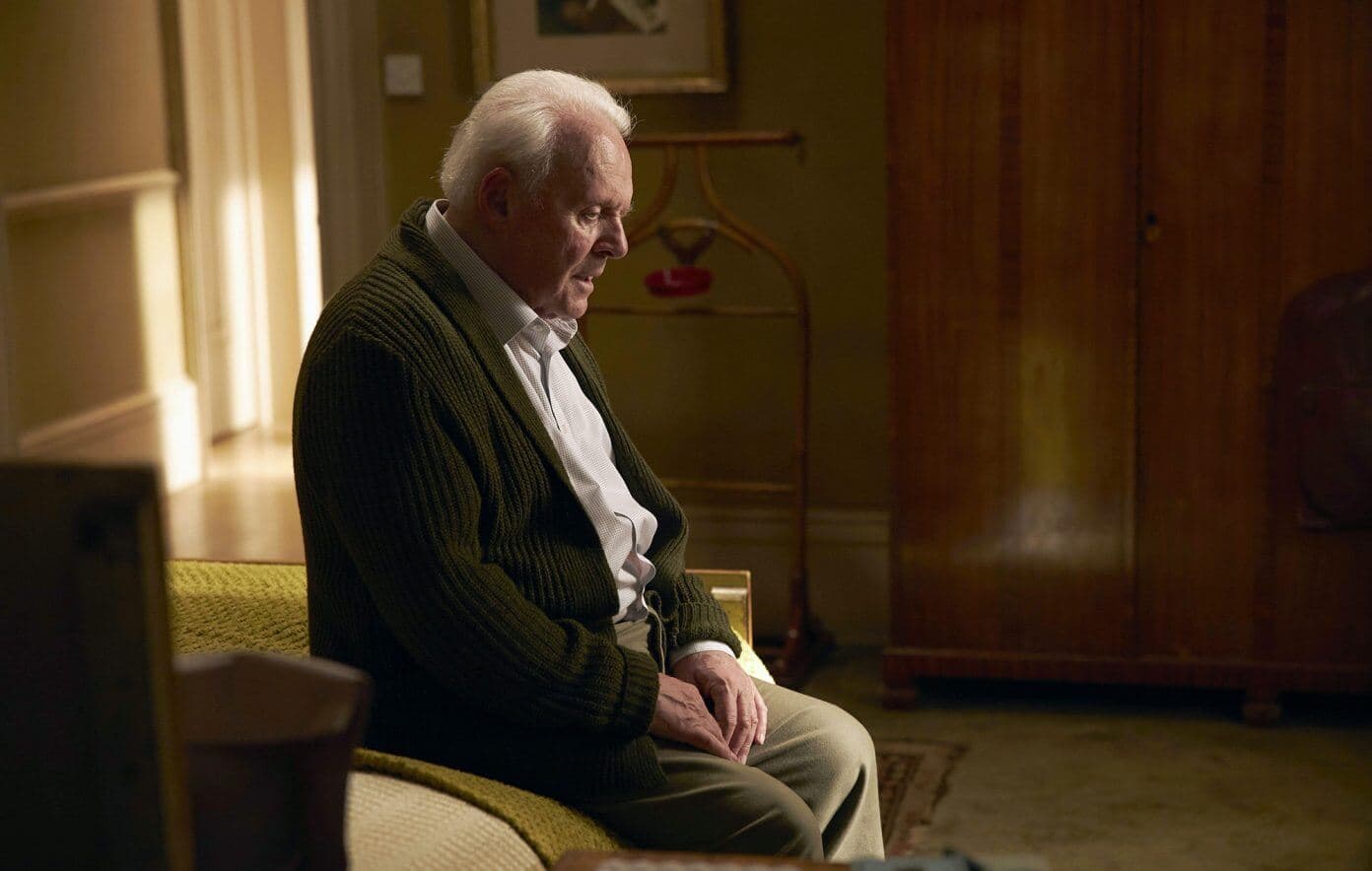 Elderly man with white hair sitting on a bed, looking pensive, in a warmly lit room with wooden furnishings. - Home Instead