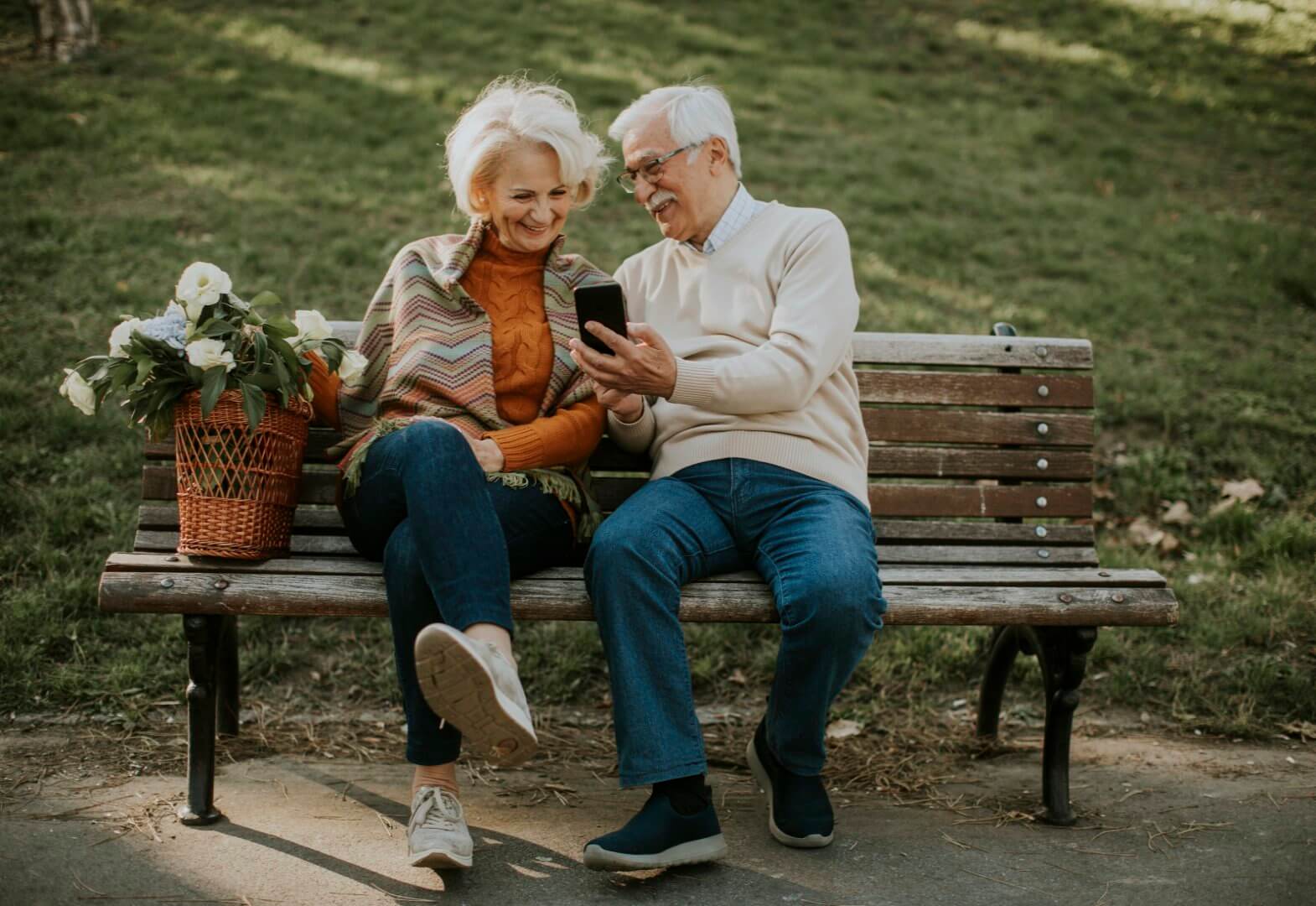 Elderly couple sitting on a park bench, smiling and looking at a smartphone, with a basket of flowers beside them. - Home Instead Southampton