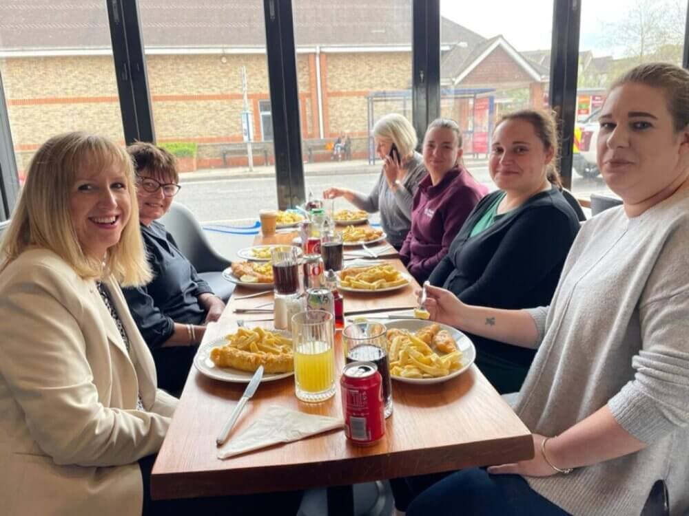 Five people sit at a table in a restaurant, eating fish and chips, smiling for the camera. - Home Instead