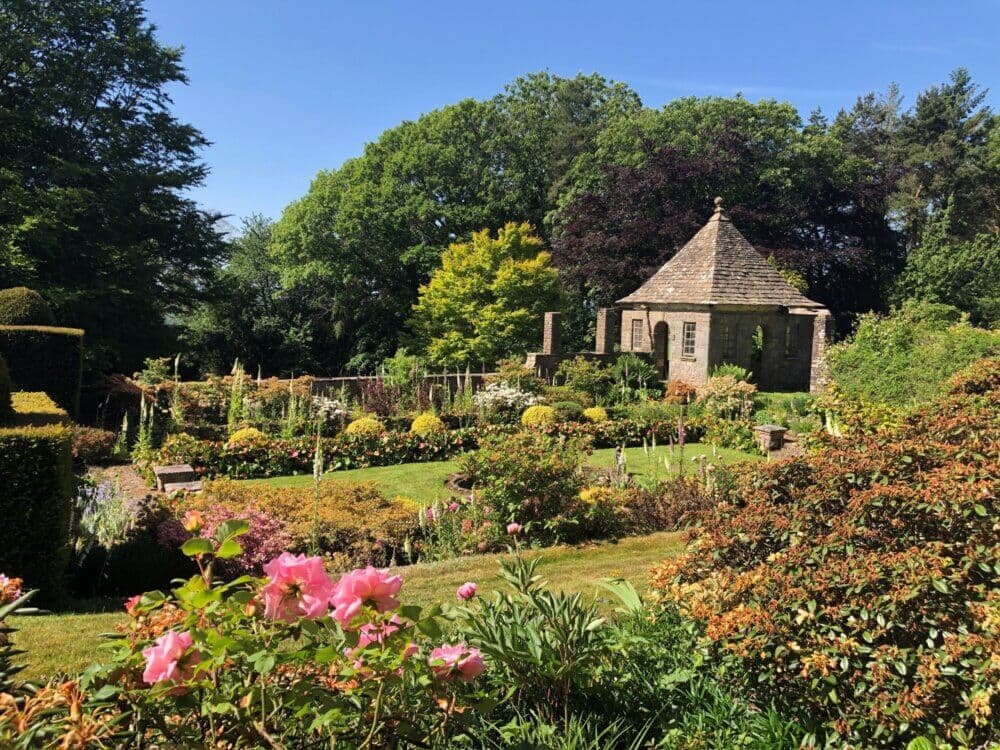 A lush garden with blooming flowers and a small stone building surrounded by trees on a sunny day. - Home Instead