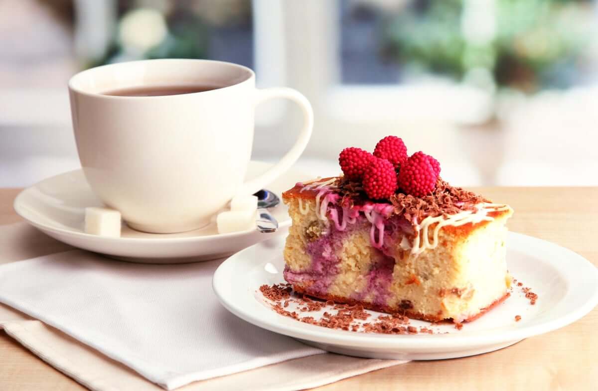 White cup of coffee next to a slice of cake topped with raspberries and chocolate shavings on a white plate. - Home Instead