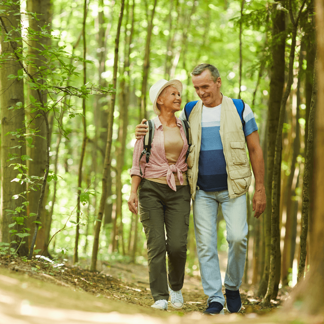 A smiling elderly couple walking arm in arm on a forest path with sunlight filtering through the trees. - Home Instead