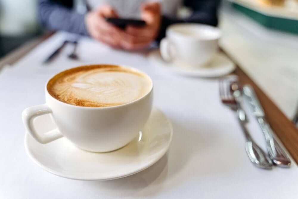 Close-up of a cup of coffee on a table with another cup and a person using a smartphone in the background. - Home Instead
