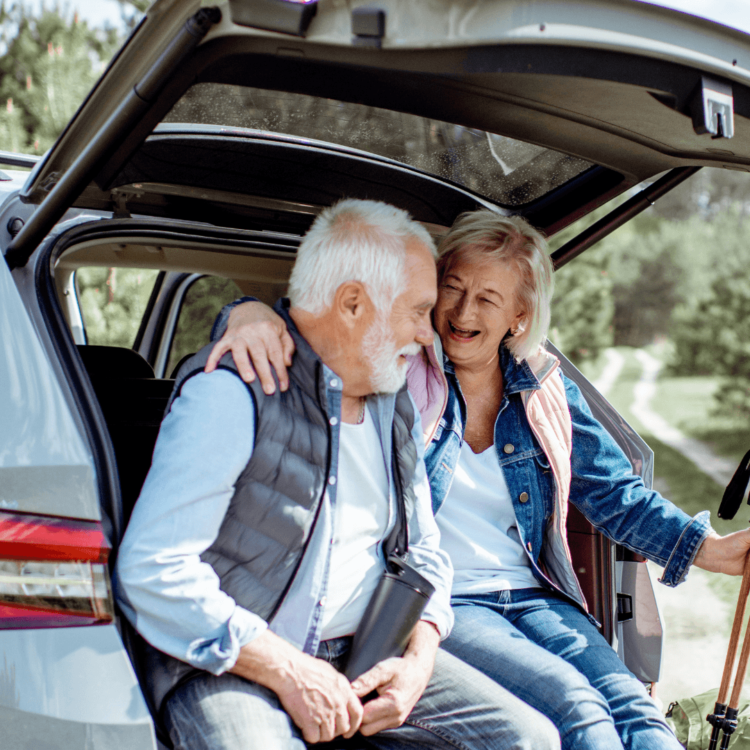 Smiling elderly couple sit in an open car trunk, embracing each other in a cheerful outdoor setting. - Home Instead