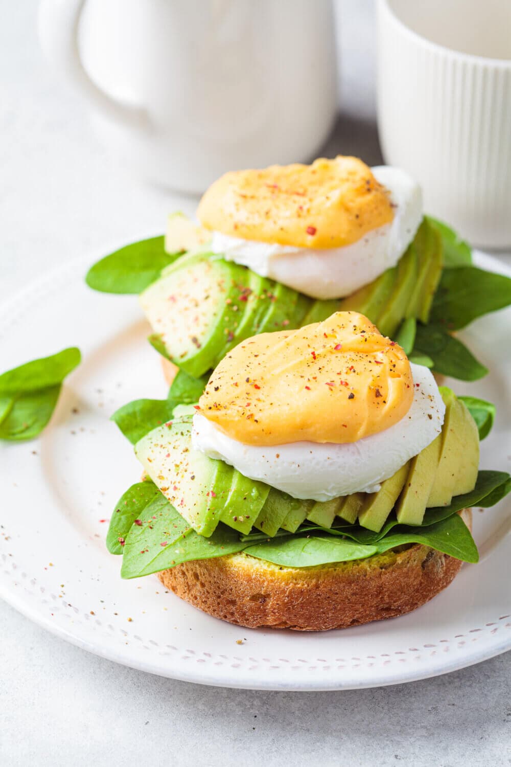Close-up of avocado toast with poached eggs, spinach, and spices on a white plate, next to a mug and pitcher. - Home Instead