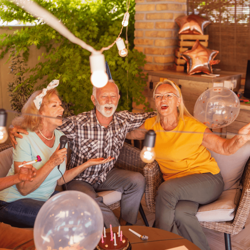 Three joyful elderly people celebrating a birthday with cake, balloons, and string lights on an outdoor patio. - Home Instead