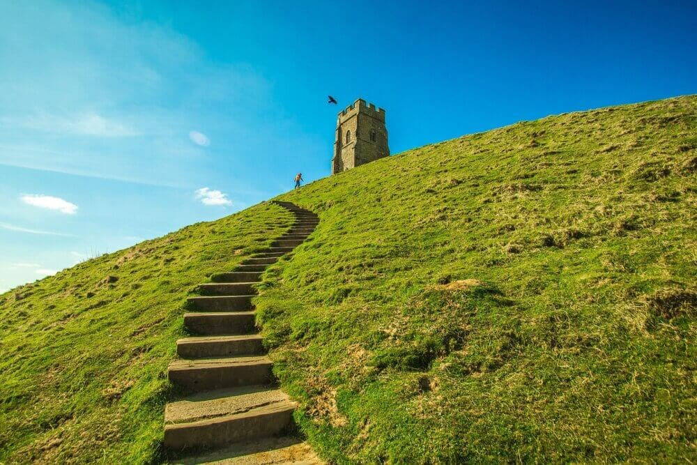 Stone steps lead up a grassy hill to a lone tower under a blue sky, with a single bird flying above. - Home Instead