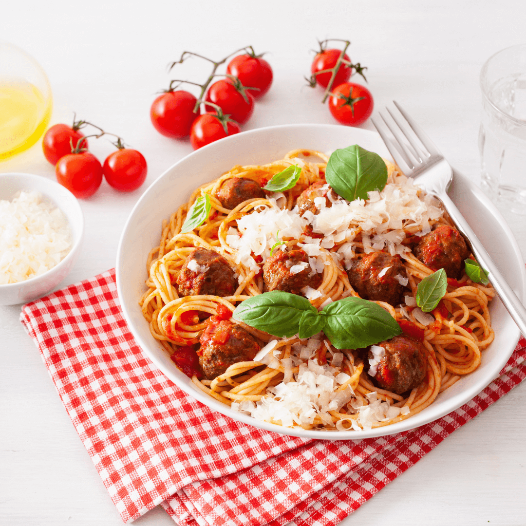 A plate of spaghetti with meatballs, topped with grated cheese and basil, on a red checkered cloth, with tomatoes on the vine nearby. - Home Instead