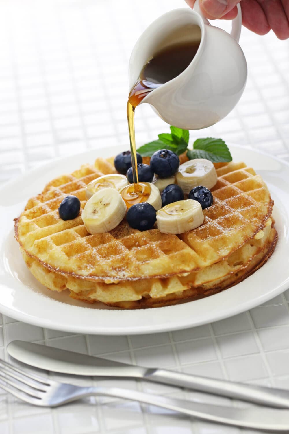 A stack of waffles topped with banana slices and blueberries, with syrup being poured over them. Silverware is next to the plate. - Home Instead