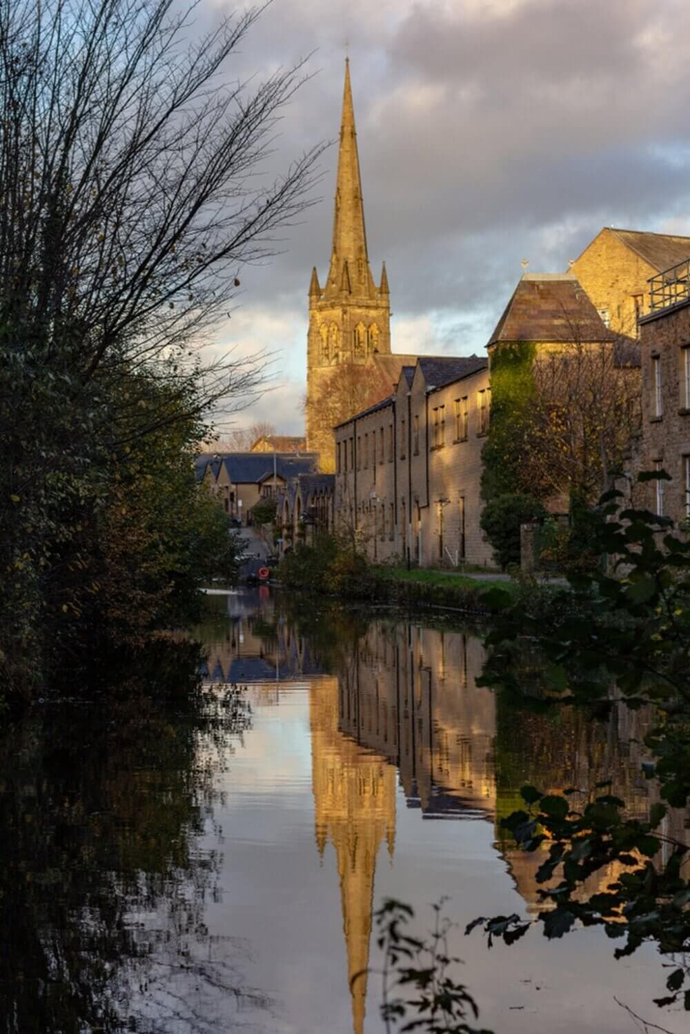 A church spire reflecting in a calm river, flanked by bare trees and stone buildings under a cloudy sky at dusk. - Home Instead