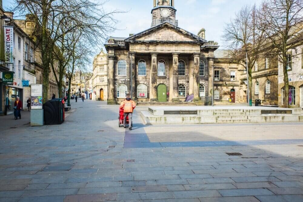 A person in an orange jacket walks with a stroller near a historic building with columns in a mostly empty plaza. - Home Instead
