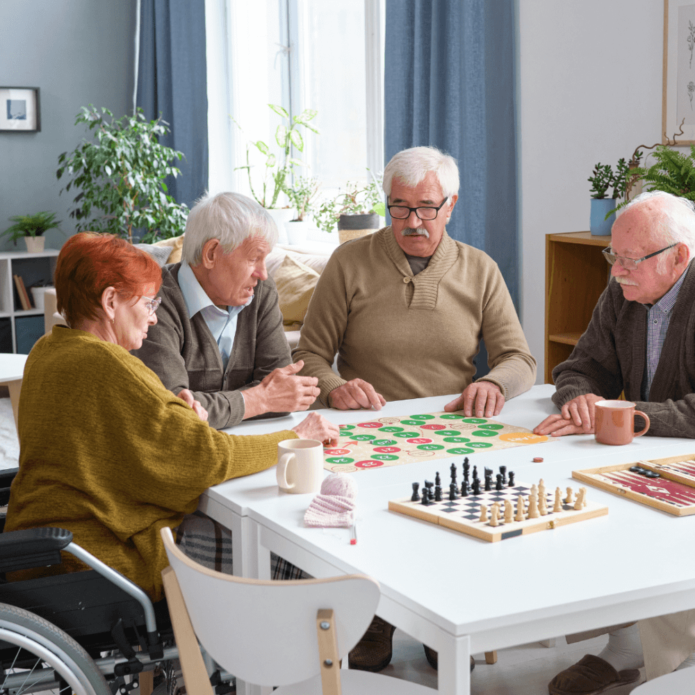 Four elderly people playing a board game together at a table, with a chessboard and plants in the background. - Home Instead