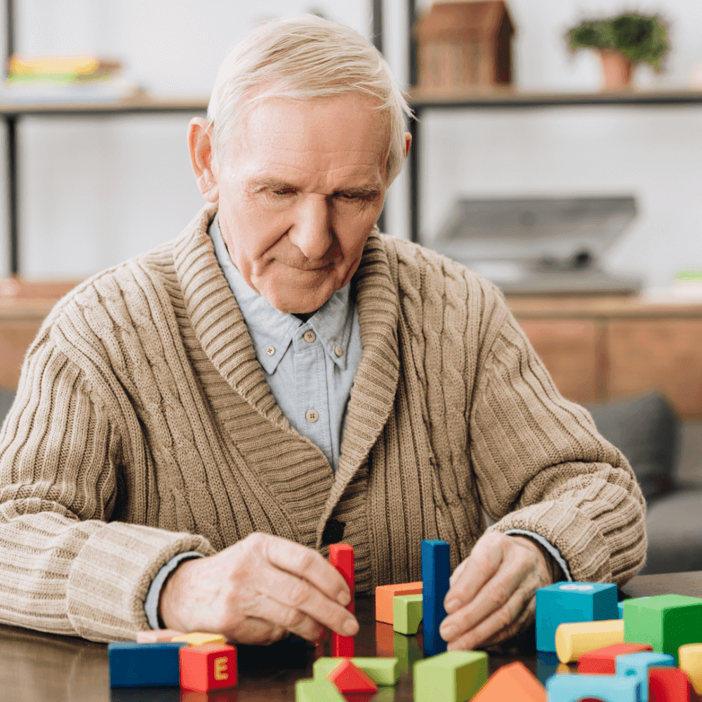 Elderly man with gray hair, wearing a brown cardigan, focused on building with colorful toy blocks on a table. - Home Instead