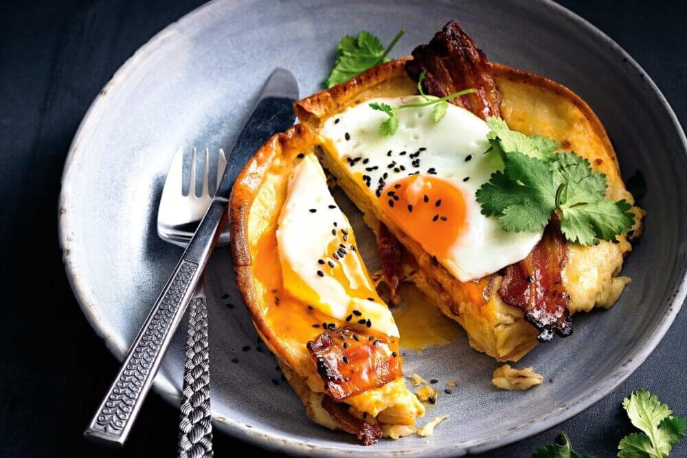 A savory dish with an egg, bacon, herbs, and black sesame seeds on a pancake, served on a plate with a fork and knife. - Home Instead