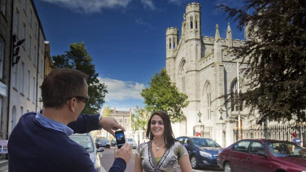A man takes a photo of a woman standing on a street with a large church building in the background on a sunny day. - Home Instead