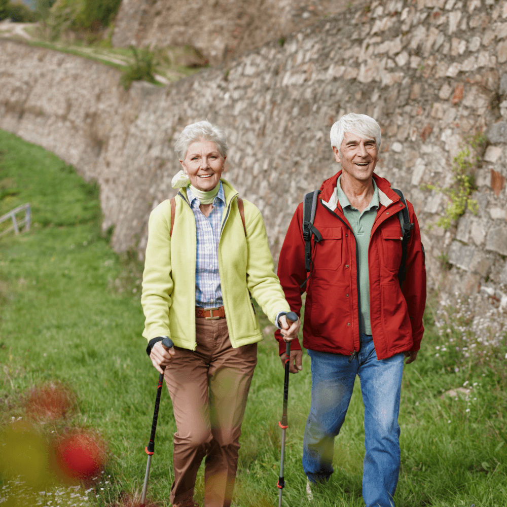An elderly couple with backpacks hiking next to a stone wall, smiling and enjoying the outdoors. - Home Instead