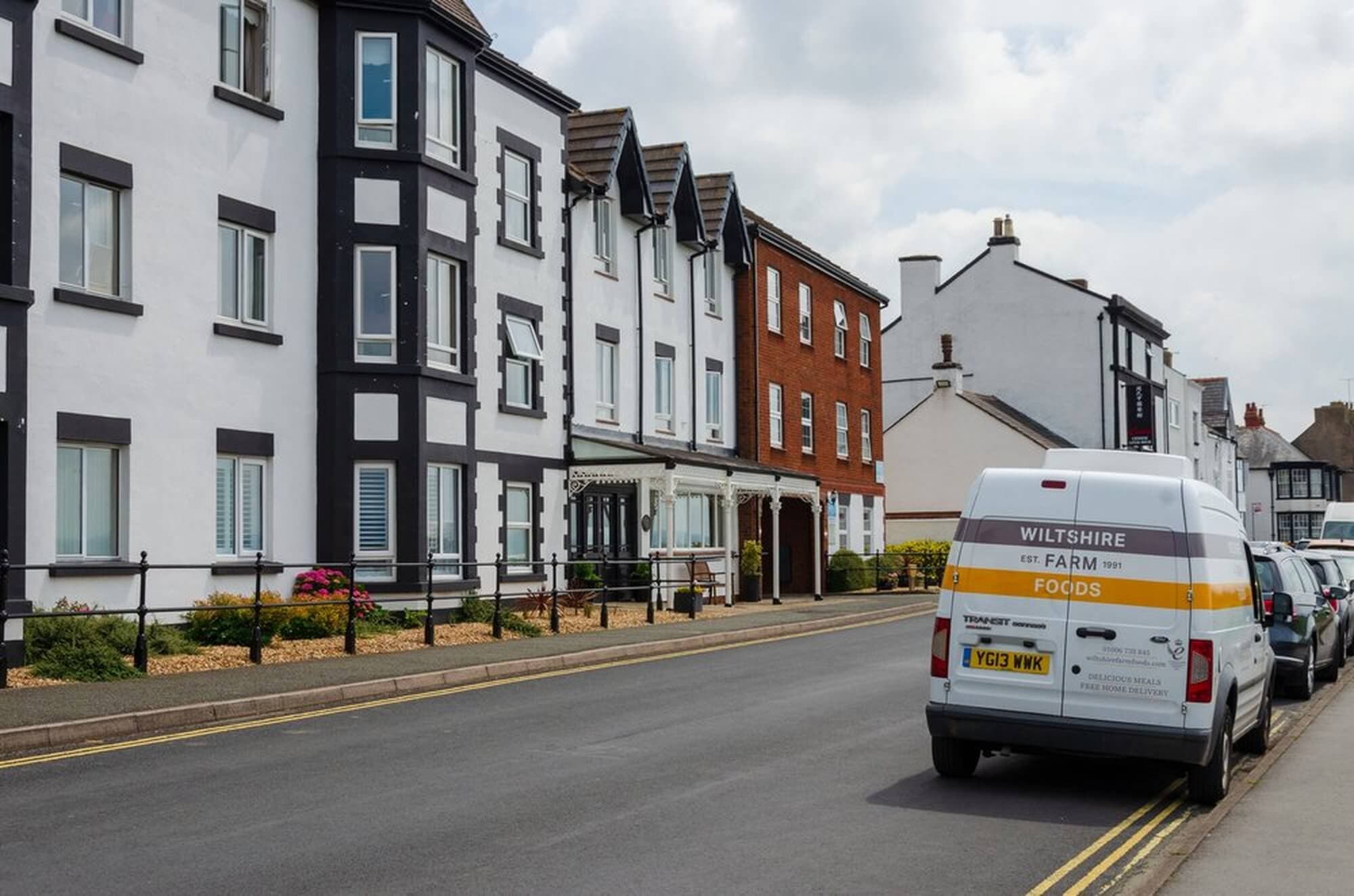 Residential street with apartment buildings and a parked white van labeled "Wiltshire Farm Foods. - Home Instead