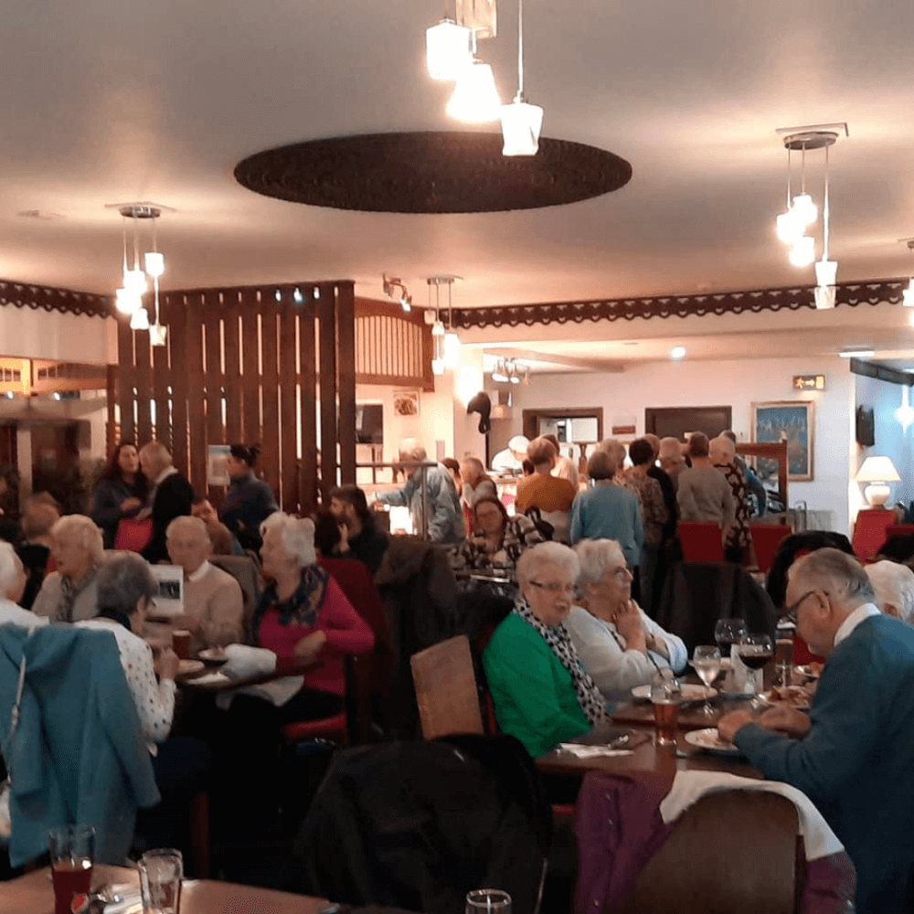 A crowded restaurant with people sitting at tables and others standing in conversation under warm lighting. - Home Instead