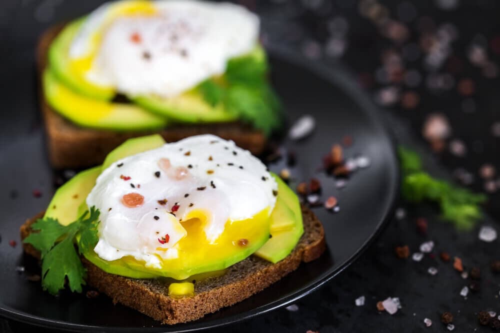 Two slices of toast with avocado and poached eggs, garnished with herbs and seasoning, on a black plate. - Home Instead